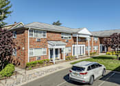 Thumbnail 5 of 13 - A silver car is parked in front of a brick building with a white awning.