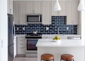 Thumbnail 54 of 82 - Studio Apartments in Beverly MA - Holmes Beverly - Modern Kitchen with White Countertops, Stainless Steel Appliances, Blue Tile Backsplash, Soft Grey Cabinetry, Large Cooking Island with Brown Stool Seating and Hanging Lights, and Plank Wood Flooring