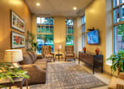 Thumbnail 12 of 22 - Liberty Gateway Apartments, Salt Lake City, Utah, lounge area with a brown couch, a chair, a table, a lamp, a plant, a rug, a television, and a painting on the wall.