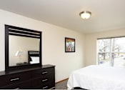 Thumbnail 9 of 12 - A bedroom with a white bed, a black dresser with a mirror, and a window with a view of trees.