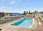 Thumbnail 3 of 24 - A swimming pool on a wooden deck with a city skyline in the background.