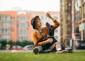 Thumbnail 20 of 20 - A woman in a yellow sweater and brown hat is sitting on the grass in front of a row of buildings.