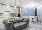 Thumbnail 1 of 14 - A grey tufted sofa sits in a kitchen with white cabinets and a marble floor.