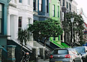 Thumbnail 5 of 14 - A street with cars parked on the side and a green tree in front of a building.