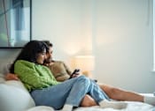 Thumbnail 5 of 54 - A man and woman are sitting on a bed, with the woman holding a remote control.