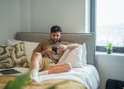 Thumbnail 17 of 54 - A man is sitting on a bed with a laptop and a phone in his hands.