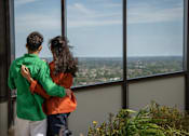 Thumbnail 37 of 54 - A couple in a green and orange jacket are standing close together and looking out a window at a cityscape.
