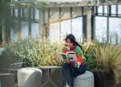 Thumbnail 39 of 54 - A woman is sitting on a grey stool reading a book in a sunny outdoor area.