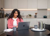 Thumbnail 14 of 54 - A woman in a pink cardigan is working on her laptop at a table with a cup of coffee and a tray of coffee cups.