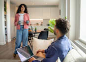 Thumbnail 9 of 54 - A woman standing in a kitchen holding a mug while a man sits on a couch using a laptop.