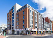Thumbnail 11 of 41 - A modern multi-story building with a mix of brick and white panels is situated on a street corner with shops in the foreground.