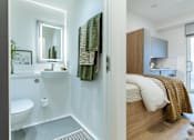 Thumbnail 9 of 17 - A bathroom with a toilet, sink, mirror, and a bedroom with a bed and a nightstand.