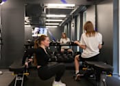 Thumbnail 27 of 51 - A woman is doing squats in a gym with another woman sitting on a bench in the background.