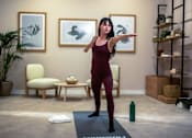 Thumbnail 44 of 85 - A woman in a maroon top and leggings is standing on a yoga mat in a room with a chair, a table, and two framed pictures on the wall.