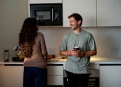 Thumbnail 83 of 85 - A man and a woman are standing in a kitchen, the man is holding a cup and they are both looking at a microwave.
