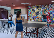 Thumbnail 20 of 49 - A woman in a blue dress is playing ping pong in a room with a colorful mural on the wall.