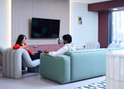 Thumbnail 24 of 49 - Two people sitting on a couch in a room with a television and a window.