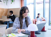 Thumbnail 15 of 49 - A person is writing in a notebook with a laptop and a lamp on the table.