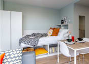 Thumbnail 26 of 51 - Side-view angle of a bed, with white bedding, a grey throw and yellow/orange pillows. Includes view of under-bed storage and a small dining table and chairs.