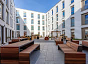 Thumbnail 17 of 48 - A modern courtyard with wooden benches and a white building in the background.