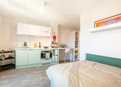 Thumbnail 5 of 21 - A bedroom with a bed, a kitchenette, and a painting on the wall.