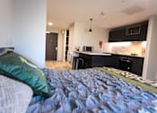 Thumbnail 10 of 23 - A bedroom with a bed and a kitchen area in the background.