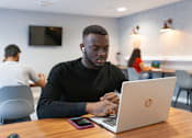 Thumbnail 31 of 51 - A man in a black shirt is using a laptop in a room with other people in the background.