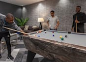 Thumbnail 21 of 51 - Three men playing pool in a room with a grey couch and a plant.