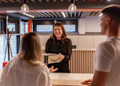 Thumbnail 36 of 51 - A woman in a black dress is holding a box with the Amazon logo on it and talking to two people.