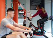 Thumbnail 8 of 51 - A man and a woman are exercising on stationary bikes in a gym.