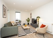 Thumbnail 15 of 23 - A living room with a grey couch, a striped rug, and a green plant.