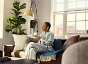 Thumbnail 17 of 24 - A woman in pajamas sits on a couch with a cup of coffee.