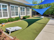 Thumbnail 23 of 61 - Outdoor grassy area with a cornhole game under triangular shade sails hanging above near a building