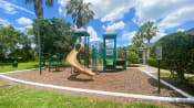 Thumbnail 5 of 11 - Green and yellow Playground set in a bed of mulch with buildings and palm tree, and bench in the background,