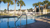 Thumbnail 10 of 15 - Community pool with sundeck, lounge chairs, with lake view surrounded by white metal fence with palm trees and building exteriors in the background