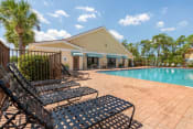 Thumbnail 6 of 24 - Leasing Office Exterior with Fenced in Swimming Pool and Sun Deck with Lounge Chairs with Palm Trees and Building Exteriors in the Background