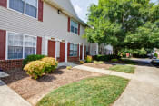 Thumbnail 4 of 20 - The apartment building exteriors are cream to tan in color with white trim and red shutters and doors. There are sidewalks leading to individual apartment doors, grass in areas between. the sidewalks, and large mature tree outside.