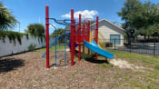 Thumbnail 5 of 31 - Red and blue playground set in a bed of mulch surrounded by a black fence with buildings and palm tree in the background