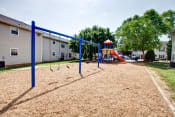Thumbnail 2 of 20 - The community playground is surrounded by mulch, grass, trees and apartment buildings. It has 4 swings, and a jungle gym area with a red slide, yellow monkey bars, and stairs.