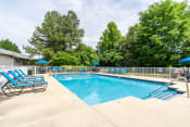 Thumbnail 1 of 20 - The swimming pool is a large rectangle surrounded by an expansive sundeck with lounge chair seating. The pool furniture is a cool blue and includes lounge chairs, three umbrella, and three round tables. The pool is enclosed by a white fence and is surrounded by large grassy areas and trees.