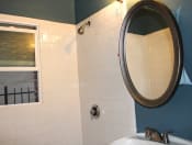 Thumbnail 13 of 28 - Bathroom in one bedroom brookmore apartment in Pasadena. Shower with white tile and silver fixtures and open window, and oval mirror above pedestal sink
