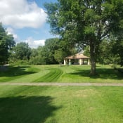 Thumbnail 11 of 54 - Large green pasture with a sidewalk across the middle. Gazebo with tables and chairs can be seen by the trees surrounding the grass. Blue and cloudy sky.