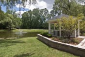 Thumbnail 9 of 16 - White gazebo to the right of photo with trees, greenery and lined with wood surround.  Green grass in the middle leading up to a pond with a fountain.  Sky and green trees in the background.