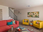 Thumbnail 9 of 13 - Living room with two yellow chairs on right wall and artwork of plant above, pink/orange couch on left wall and coffee table in center of room.  Stairs to upper level in background.
