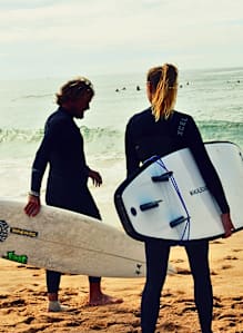 Two people in wetsuits holding surfboards on a beach.