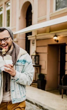 Two men walking down the street, laughing and eating noodles from takeout boxes.
