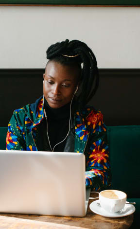 Woman sitting at a table with a laptop, coffee, and earphones on.