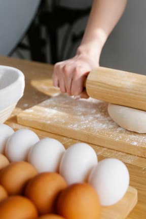 A person is rolling out dough on a wooden surface with eggs nearby.