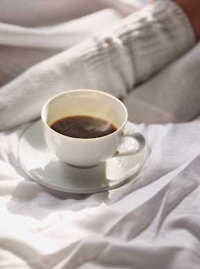 A cup of coffee sits on a saucer with a person's arm and a white blanket in the background.