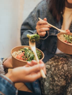 two women eating a bowl of food with chopsticks